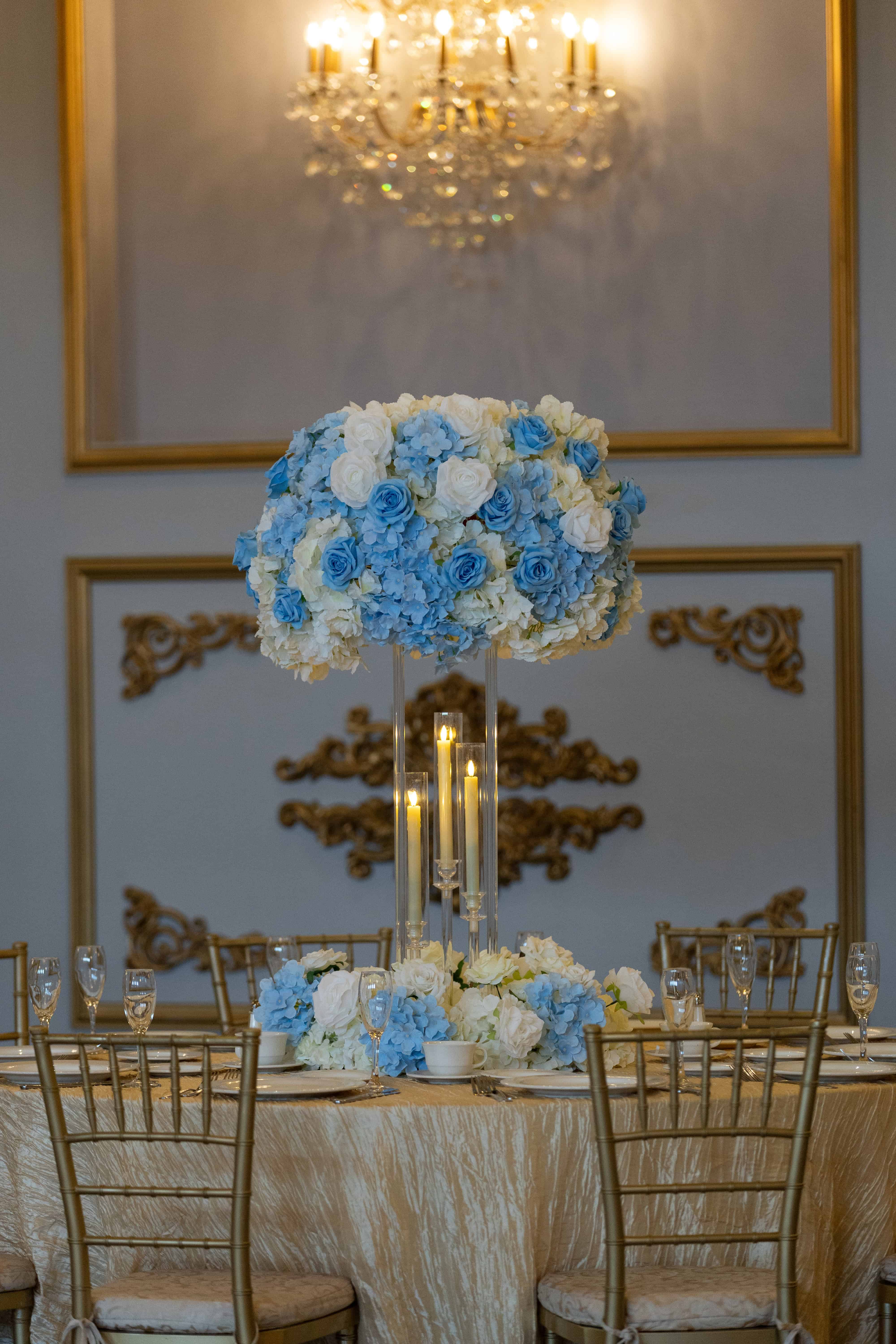 Elegant wedding centerpiece with blue and white roses, gold accents, and candlelit glow for event rentals.